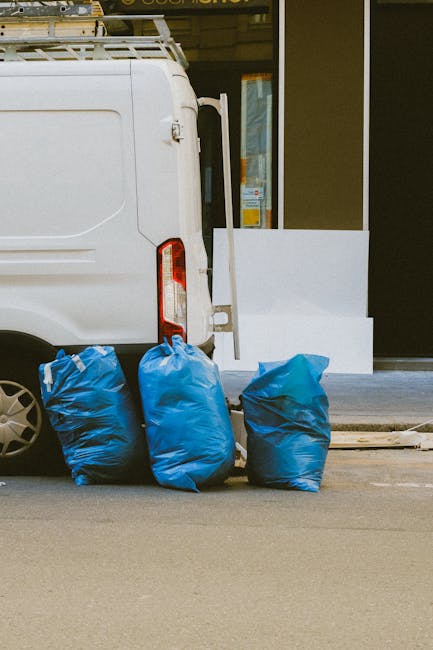 A close-up view of three large blue rubbish bags positioned on the pavement beside the rear of a white service van, which is parked in front of a building with a glass door and window. The bags are made of thick plastic with a slightly crumpled texture and are filled with waste, causing them to appear bulky and rounded. The van's rear door is closed, with part of a ladder rack visible on top, indicating a commercial or waste collection vehicle. The scene suggests an outdoor rubbish removal process, possibly carried out by waste collection services like Waste Collection Earls Court, with the bags awaiting disposal or transfer for private disposal or alternative waste handling. The environment appears urban, with a concrete sidewalk and a storefront background visible through the glass, and the lighting is natural, implying daytime conditions.