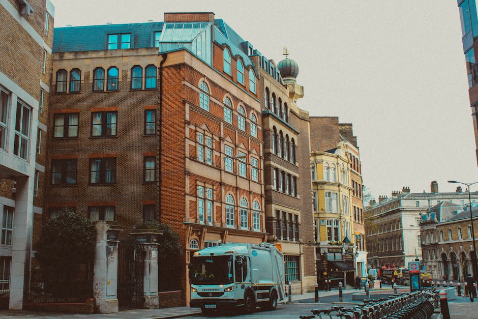 A waste collection vehicle with a large, open rear loading compartment is positioned on a narrow cobblestone street, adjacent to a row of older, multi-story buildings with weathered facades. The vehicle's exterior is white, with visible signs of rust and dirt near the rear opening, and features mechanical components such as hydraulic arms and collection mechanisms at the back. A worker dressed in a blue uniform and an orange high-visibility vest is standing beside the truck, manually depositing waste into a blue plastic bin or container. The background includes parked cars, including a black vehicle directly behind the waste truck, and urban signage indicating parking restrictions. Natural daylight illuminates the scene, highlighting the textures of the cobblestones, the weathered building surfaces, and the metallic and plastic elements of the waste collection equipment. Waste Collection Earls Court specializes in private rubbish removal, often providing alternative waste handling services outside of local authority collection schedules, as evidenced by the operational scene depicted.
