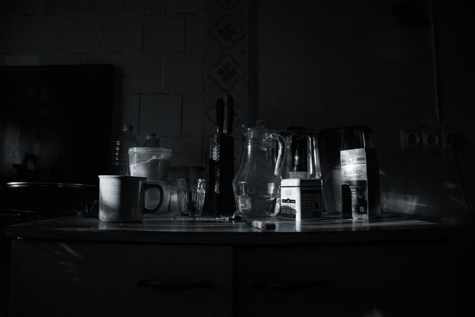 A dimly lit photograph of a kitchen countertop featuring various items arranged in a cluttered manner. Prominently, there is a white ceramic mug with a handle, situated towards the left side of the surface. Next to the mug are several glassware pieces, including a tall glass and a smaller tumbler, all with clear, smooth surfaces. Behind the glassware, there is a plastic container with a lid, slightly opaque, and a plastic bottle. To the right, a clear glass pitcher with a handle occupies the central position, partially filled with water. Further to the right, a black and silver electric kettle with a glossy finish stands, with a visible white label indicating it's an Earl Grey tea kettle. Next to it, a small white box and a carton with a spout are present, along with kitchen utensils like knives housed in a knife block and a pair of tongs. The background reveals ceramic tiles in light grey tones, some with faint decorative patterns, with an embedded electrical outlet visible on the right. The surface appears to be a kitchen countertop or table, and the lighting suggests an indoor environment with natural or low ambient light, emphasizing the reflective surfaces of the glassware and appliances. Waste collection and disposal are contextual factors, though not directly depicted, this arrangement relates to typical household waste items or prep clutter often managed through private or council rubbish removal services.