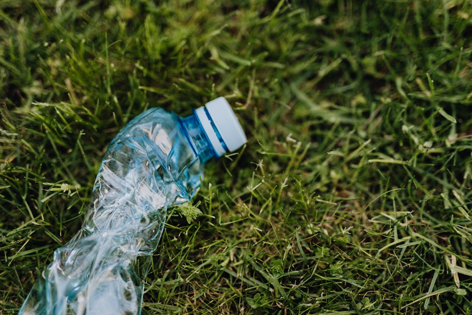 A crumpled, transparent plastic water bottle with a white and blue screw-on cap lies on a patch of uneven green grass, with blades of grass of varying lengths and textures, some appearing freshly cut and others longer. The grass covers the ground densely, and the bottle is positioned in the foreground, slightly tilted to one side, with its crinkled surface reflecting light. In the background, the grassy area continues, with no visible structures or other objects. This scene highlights common litter found in outdoor environments, relevant to discussions on rubbish removal or private waste disposal services such as those offered by Waste Collection Earls Court, particularly for managing loose waste like plastic bottles in the context of alternative waste handling methods.