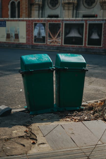 Two green plastic bins with closed, slightly domed lids positioned side by side on the edge of a paved sidewalk, close to the curb. The bins are made of textured, durable plastic with black hinges and appear to be for general waste collection. They are situated outdoors in an urban setting, with a concrete pavement in the foreground that shows some soil and debris around the base. In the background, there is a street paved with asphalt and a brick building with large, framed windows, some covered with posters or advertisements. The scene is lit by natural daylight, casting soft shadows, and the overall setting suggests the bins are for private waste disposal, possibly part of a local or independent rubbish collection service, consistent with alternatives to municipal rubbish collection services.