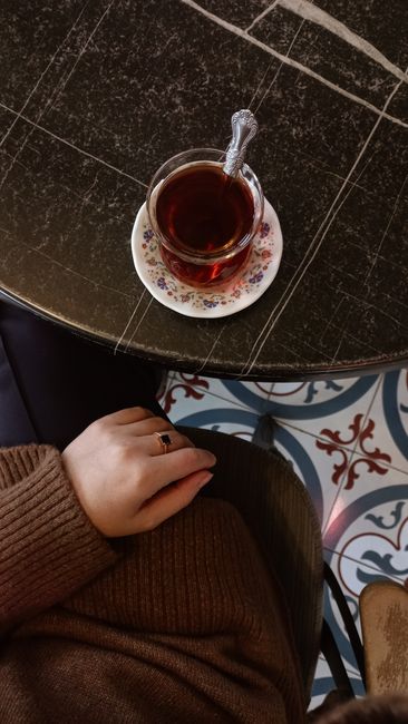 A close-up view of a white ceramic cup filled with hot tea, placed on a metallic saucer with a small metallic spoon inside. Above the cup, a used tea bag with a dark brown, slightly damp fabric is suspended in mid-air, dripping a small droplet of tea. The background is softly blurred with neutral and warm tones, suggesting a cozy indoor setting. The lighting highlights the glossy finish of the cup and saucer, and the texture of the tea bag, which appears slightly wrinkled and wet. This scene relates to household waste disposal, as the used tea bag represents organic waste that may be handled via private rubbish collection or on-site clearance services, such as those offered by Waste Collection Earls Court.