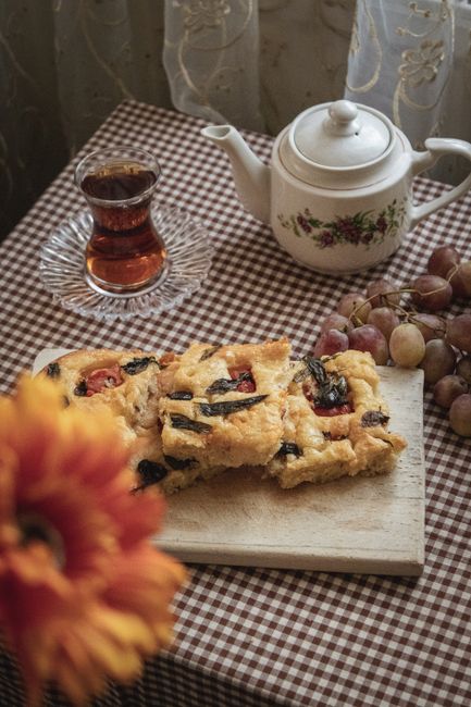 The image depicts a close-up view of a table set with a checkered tablecloth, featuring a layered fruit and sponge cake with visible berries and dark fruit pieces on a small wooden cutting board in the foreground. Behind the cake, there is a white ceramic teapot with a floral design, a matching lid, and a curved spout positioned slightly to the right. To the left of the teapot, a clear glass cup filled with dark tea sits on a small glass saucer with a textured edge. In the background, a cluster of grapes in shades of purple and green rests on the table, partially obscured by the foreground objects, with a sheer curtain featuring subtle floral embroidery hanging behind the table, allowing soft natural light to illuminate the scene. The overall atmosphere is cozy and domestic, with a hint of rustic charm, suitable for describing a setting related to food, tea, and casual home entertaining.