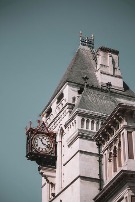 A close-up view of a historic clock tower attached to a tall, white stone building, featuring ornate architectural details and decorative battlements at the top. The clock face, surrounded by a dark frame, displays black Roman numerals and is mounted on a projecting section of the tower, with the time clearly visible. The building's facade showcases a combination of smooth and textured stone surfaces, with pilasters, arched windows, and cornices highlighting classical architectural elements. The slate roof of the tower is steeply pitched, with a small decorative metal spire or lightning rod at the peak. The sky behind the tower is clear and muted, providing a neutral backdrop that emphasizes the structure's historical and architectural details. This scene, often associated with London landmarks, subtly relates to independent collection and on-site clearance services provided by Waste Collection Earls Court, aligning with the concept of alternative waste handling in prominent urban environments.
