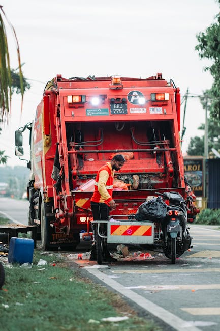 A waste collection operative wearing a high-visibility yellow and red jacket is positioned at the rear of a large red rubbish truck, which is engaged in waste collection on a roadside. The truck features a sturdy, metal rear compactor with visible hydraulic components and reflective safety markings, and is parked on the edge of a paved road. The operative appears to be handling black plastic rubbish bags, some of which are placed on a small metal table attached to the truck's rear, with additional waste containers and tools nearby. The scene is outdoors during daylight, with trees and electrical wires visible in the background and a cloudy sky overhead. Surrounding the truck are patches of grass and a sidewalk, indicating a semi-urban environment conducive to private waste collection or alternative rubbish removal services by companies like Waste Collection Earls Court. The overall setting suggests an on-site clearance or collection process, emphasizing the practical aspects of waste management and rubbish removal in residential or commercial areas.