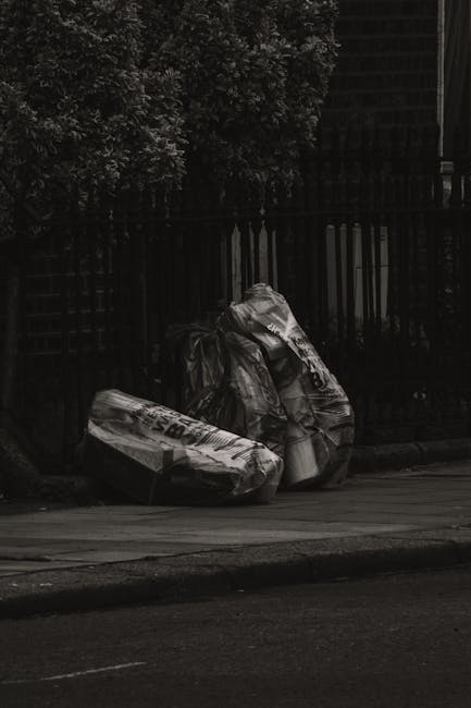 Two large black garbage bags filled with waste are placed on the sidewalk near a wooden fence and a shrub, with the bags appearing crumpled and tied at the top. The bags are positioned close to each other, leaning slightly against the fence, which consists of vertical wooden slats painted black. The environment appears to be an outdoor urban setting, with a paved road visible in the foreground and a roadway curb. The lighting suggests a somewhat overcast or shaded area, with minimal natural light illuminating the scene. This image, associated with waste collection services, illustrates private rubbish disposal or on-site clearance, which Waste Collection Earls Court could facilitate for bulky rubbish removal in the area, supporting alternative waste handling outside of standard council collection.